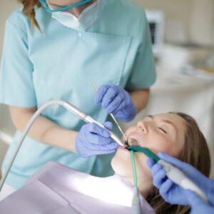 A dental hygienist, wearing a mask and gloves, uses a tool to clean a patient's teeth. The patient is lying in the dental chair with their mouth open.
