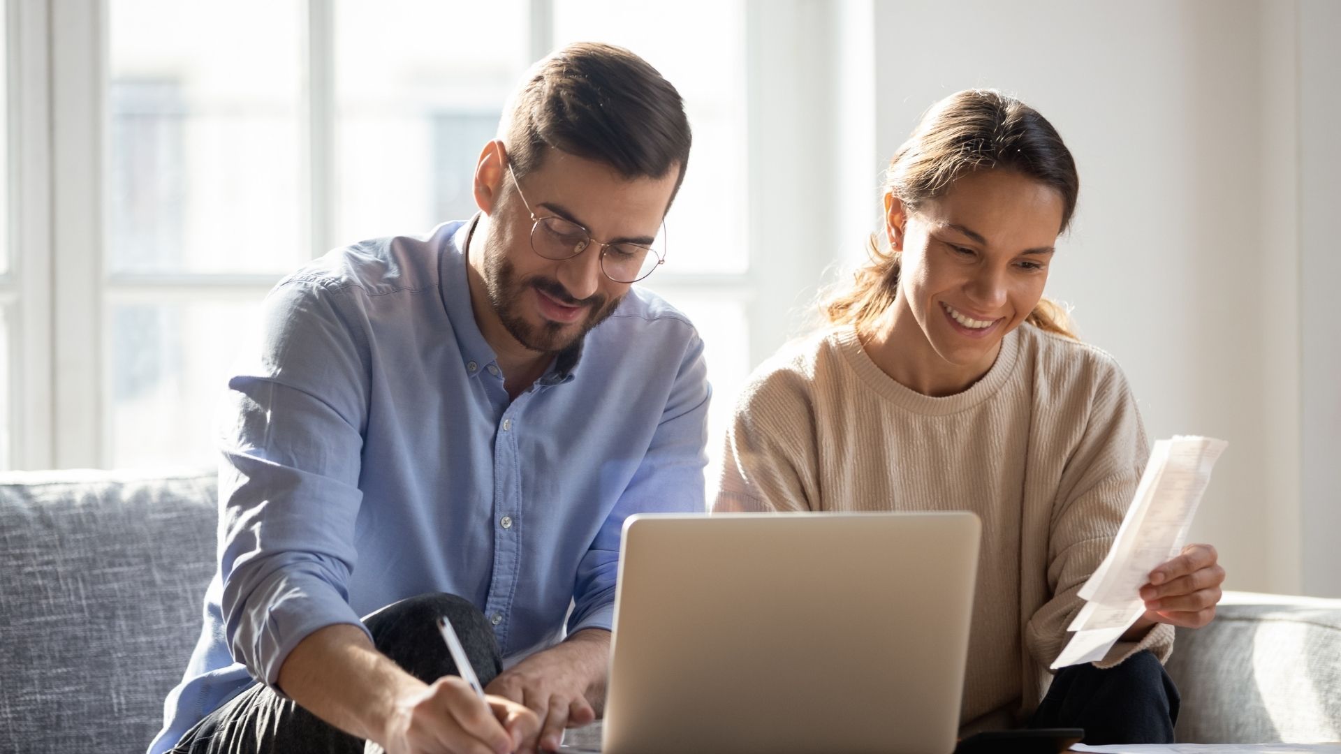 Diverse couple smiling while reviewing paperwork and calculating costs on a laptop by a sunny window; represents financial planning and affordability.