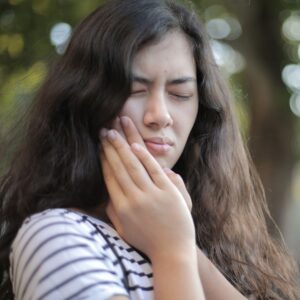 Young woman outdoors holding her cheek with both hands, clearly indicating discomfort and tooth pain.