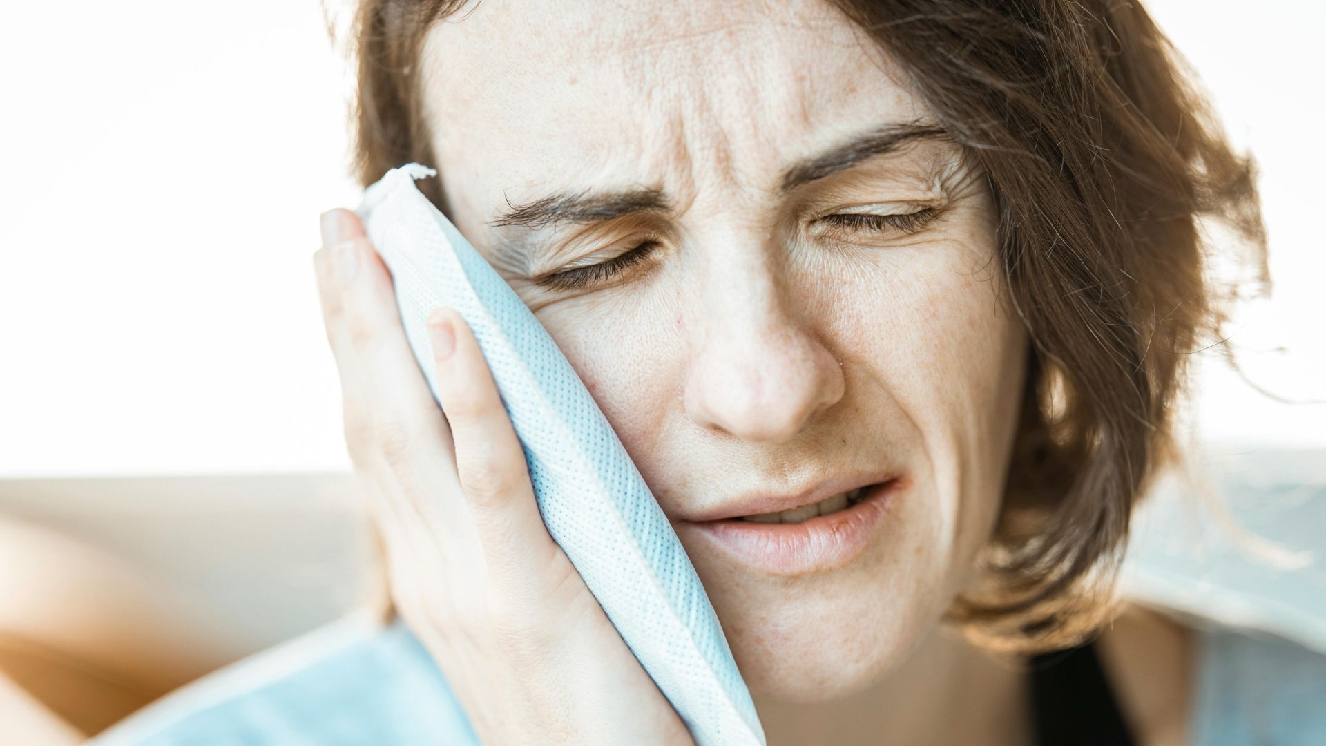 Close-up of a distressed woman with her eyes closed, holding a cold compress or ice pack to her painful cheek, illustrating severe tooth pain.